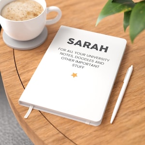 May include: A white notebook with a grey bookmark ribbon featuring the text SARAH FOR ALL YOUR UNIVERSITY NOTES, DOODLES AND OTHER IMPORTANT STUFF and a small yellow star icon, accompanied by a white pen resting on a wooden surface.