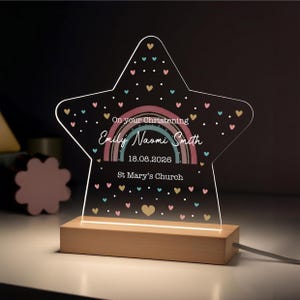 May include: A star-shaped night light with a wooden base. The clear acrylic front features a rainbow design, hearts, and the text "On your Christening" with a name, date, and church name. The light is plugged in.