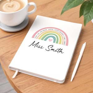 May include: A white notebook featuring a rainbow design with the words "Teach Love Encourage Inspire Listen Motivate Learn Connect". The name "Miss Smith" is written below the rainbow. A white pen and a cup of coffee are also visible.