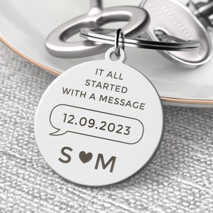 May include: A circular silver-toned metal keyring engraved with the text IT ALL STARTED WITH A MESSAGE, the date 12.09.2023 inside a speech bubble graphic, and the letters S and M separated by a heart symbol, attached to a metal split ring.