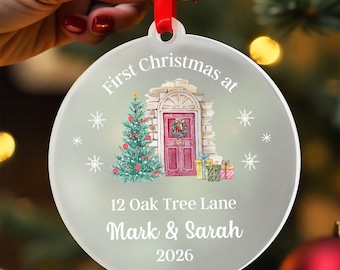 Peut inclure: Une décoration de Noël ronde en verre dépoli avec un ruban rouge. Le motif présente une illustration aquarelle d'une entrée de maison avec une porte rouge, un sapin décoré et des cadeaux. Le texte indique : First Christmas at 12 Oak Tree Lane Mark & Sarah 2026.