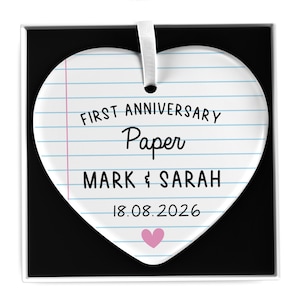 Puede incluir: Adorno en forma de coraz&oacute;n con dise&ntilde;o de papel rayado. El texto dice "FIRST ANNIVERSARY Paper MARK & SARAH 18.08.2026" con un coraz&oacute;n rosa en la parte inferior. Se adjunta una cinta blanca para colgar.