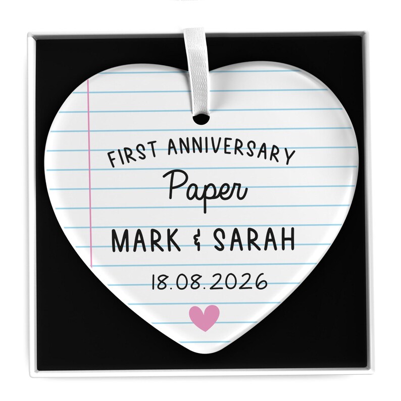 Puede incluir: Adorno en forma de coraz&oacute;n con dise&ntilde;o de papel rayado. El texto dice "FIRST ANNIVERSARY Paper MARK & SARAH 18.08.2026" con un coraz&oacute;n rosa en la parte inferior. Se adjunta una cinta blanca para colgar.