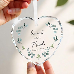 May include: A clear, heart-shaped crystal ornament with a white ribbon. The ornament features the names "Sarah AND Mark" and the date "18.08.2026" surrounded by a watercolor-style eucalyptus wreath. The ornament is held by a person.
