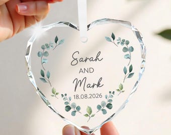 May include: A clear, heart-shaped crystal ornament with a white ribbon. The ornament features the names "Sarah AND Mark" and the date "18.08.2026" surrounded by a watercolor-style eucalyptus wreath. The ornament is held by a person.