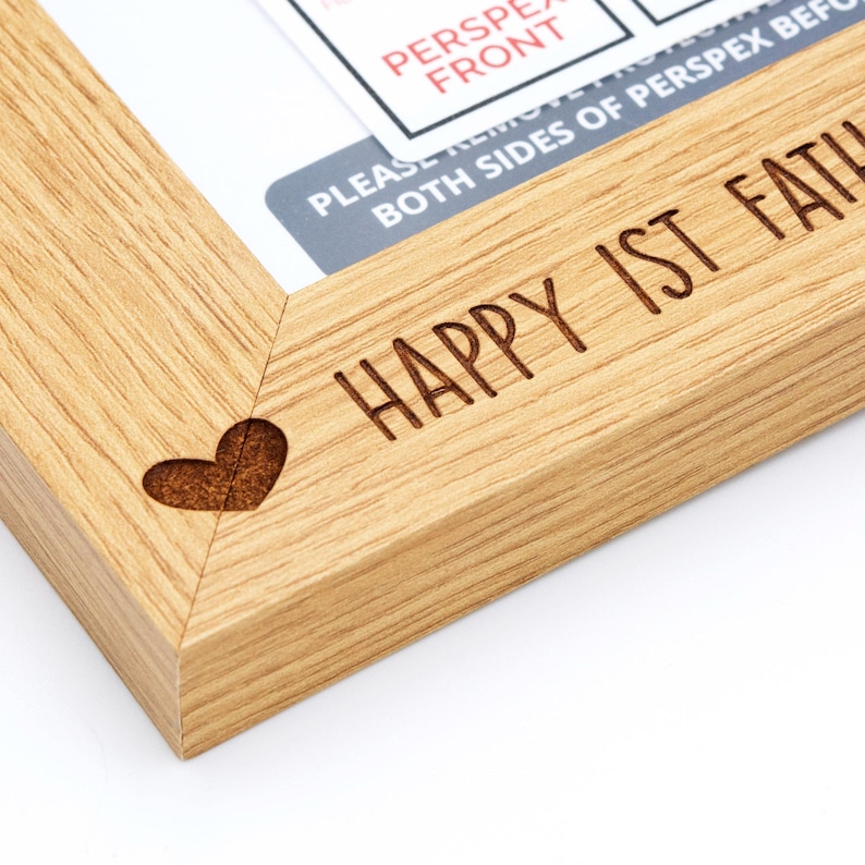 May include: A light-colored wooden picture frame with the words "HAPPY 1ST FATHER'S DAY" engraved on the frame. A small heart is carved into the corner. The frame has a natural wood grain.