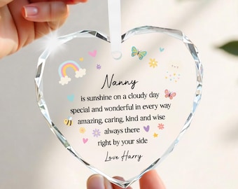 Könnte beinhalten: Herzförmiger, klarer Glasschmuck mit weißem Band. Der Schmuck trägt das Wort "Nanny" und eine Botschaft mit farbenfrohen Illustrationen eines Regenbogens, Schmetterlingen und Blumen. Die Nachricht ist mit "Love Harry" signiert.