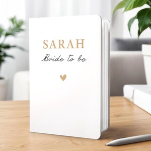 Może przedstawiać: Biały notatnik z napisem "SARAH" złotymi literami i frazą "Bride to be" czcionką. Małe złote serce znajduje się pod tekstem. Notatnik leży na drewnianej powierzchni.