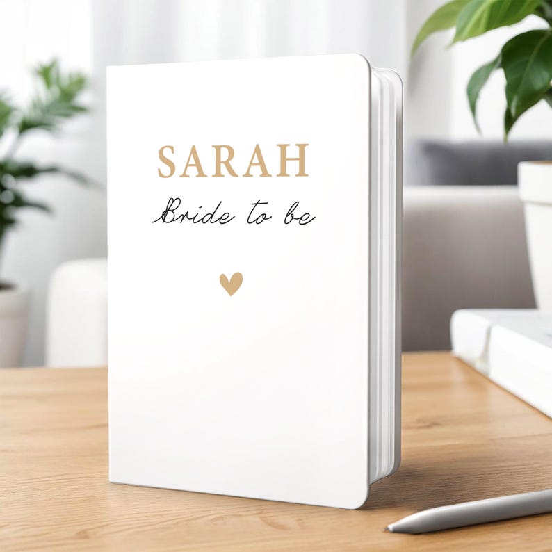Może przedstawiać: Biały notatnik z napisem "SARAH" złotymi literami i frazą "Bride to be" czcionką. Małe złote serce znajduje się pod tekstem. Notatnik leży na drewnianej powierzchni.
