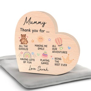 K&ouml;nnte beinhalten: Ein herzf&ouml;rmiges Holzschild mit dem Wort "Mummy" in Schreibschrift. Das Schild zeigt Illustrationen eines B&auml;ren, einer Sonne, eines Hei&szlig;luftballons, eines Regenbogens und einer Krone. Der Text enth&auml;lt "Thank you for..." und "Love Sarah."