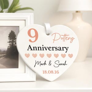 May include: A white heart-shaped ceramic plaque with a white ribbon. It displays the text "9 Pottery Anniversary" in coral and black, the names "Mark & Sarah", and the date "18.08.16". Decorative hearts are also present.
