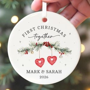 Puede incluir: Un adorno redondo de cerámica blanca con el texto FIRST CHRISTMAS together y MARK & SARAH 2026. El diseño incluye una ilustración en acuarela de ramas de pino con bayas rojas y dos adornos colgantes en forma de corazón rojo.
