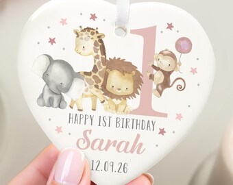 May include: Heart-shaped ceramic ornament with a white background, featuring a giraffe, lion, elephant, and monkey. The ornament has the text "HAPPY 1ST BIRTHDAY Sarah 12.09.26" in pink. A white ribbon is attached for hanging.
