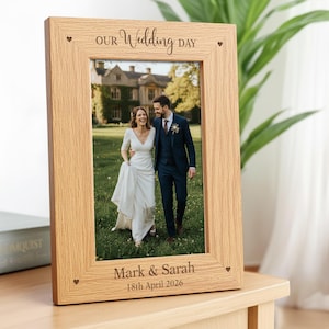 May include: A rectangular light brown wooden picture frame featuring engraved text that reads Our Wedding Day at the top and Mark & Sarah 18th April 2026 at the bottom. Small heart icons are etched into each of the four corners of the frame.
