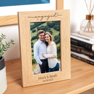 May include: A light-colored wooden picture frame with the word "engaged" in script at the top. The frame holds a photo of a couple and is engraved with the names "Mark & Sarah" and the date "12.08.2025".