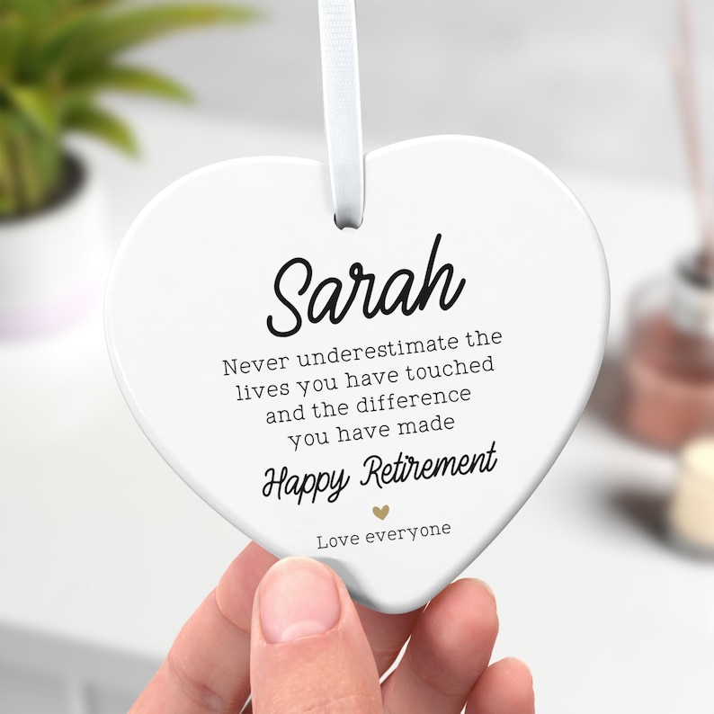 Op de afbeelding: Een wit hartvormig ornament met een wit lint. Het ornament heeft de naam "Sarah" in zwart script, samen met de tekst "Never underestimate the lives you have touched...Happy Retirement...Love everyone."