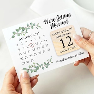 May include: A wedding save-the-date card with the text "We're Getting Married." The card shows a calendar for August 2027, with the 11th circled. A wooden square indicates the wedding date as August 12, 2027. Eucalyptus sprigs decorate the card.