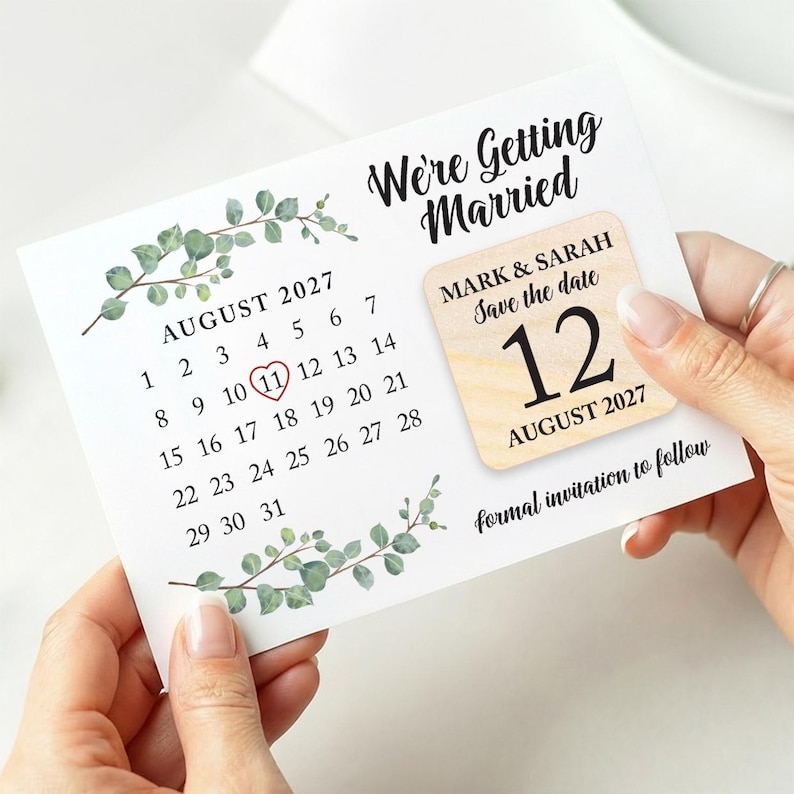 May include: A wedding save-the-date card with the text "We're Getting Married." The card shows a calendar for August 2027, with the 11th circled. A wooden square indicates the wedding date as August 12, 2027. Eucalyptus sprigs decorate the card.