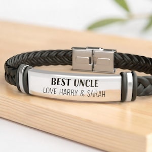 May include: Black braided leather bracelet with a silver metal plate. The plate is engraved with the words "BEST UNCLE LOVE HARRY & SARAH". The bracelet has a stainless steel clasp and is on a wooden surface.