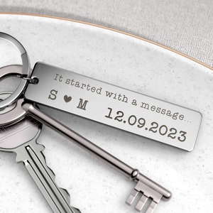 May include: A silver rectangular keychain with the text "It started with a message..." and the date 12.09.2023. The keychain also features the initials "S ❤️ M". Two keys are visible next to the keychain.