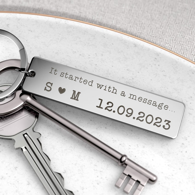 May include: A silver rectangular keychain with the text "It started with a message..." and the date 12.09.2023. The keychain also features the initials "S ❤️ M". Two keys are visible next to the keychain.