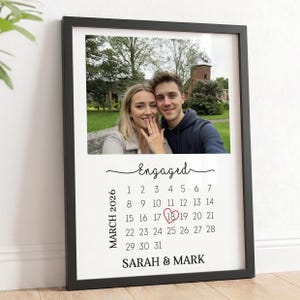 May include: Black framed print with a photograph of a couple, above a calendar. The word "Engaged" is written above the calendar, which highlights March 18, 2026. The names "SARAH & MARK" are at the bottom.