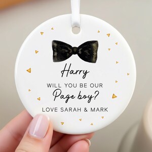 Puede incluir: Un adorno de cerámica blanca con un lazo negro y el texto "Harry, ¿quieres ser nuestro Page boy? Love Sarah & Mark." Detalles de triángulos dorados adornan el borde. Una cinta blanca está adjunta para colgar.
