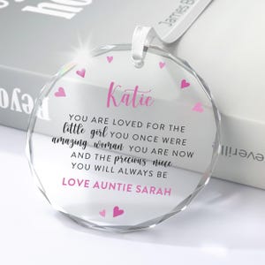 Puede incluir: Adorno de cristal transparente y redondo con una cinta blanca. El adorno presenta el nombre "Katie" en escritura rosa, un mensaje cari&ntilde;oso y acentos de coraz&oacute;n rosa. El texto dice "Love Auntie Sarah".