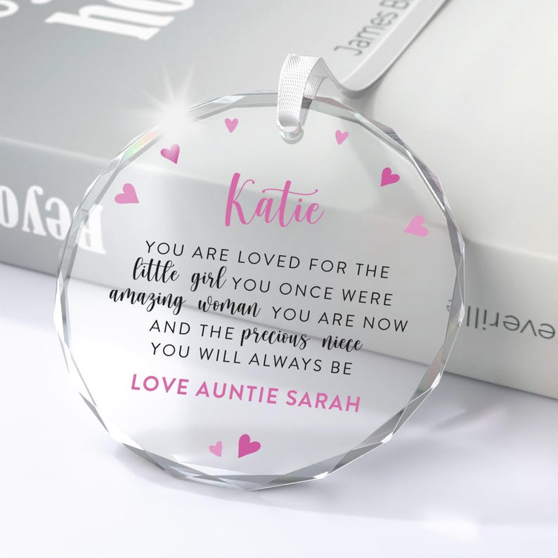 Puede incluir: Adorno de cristal transparente y redondo con una cinta blanca. El adorno presenta el nombre "Katie" en escritura rosa, un mensaje cari&ntilde;oso y acentos de coraz&oacute;n rosa. El texto dice "Love Auntie Sarah".