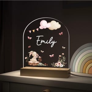 May include: A night light with a wooden base and an arched acrylic panel. The panel features a bunny illustration, the name "Emily", pink butterflies, clouds, and floral accents. The light is designed for a nursery or child's room.