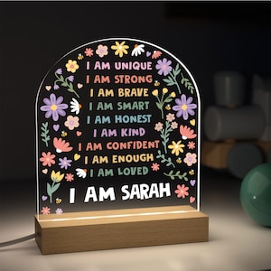May include: A decorative LED night light with a wooden base. The clear acrylic panel features colorful floral designs and the text "I AM UNIQUE, I AM STRONG, I AM BRAVE, I AM SMART, I AM HONEST, I AM KIND, I AM CONFIDENT, I AM ENOUGH, I AM LOVED, I AM SARAH."