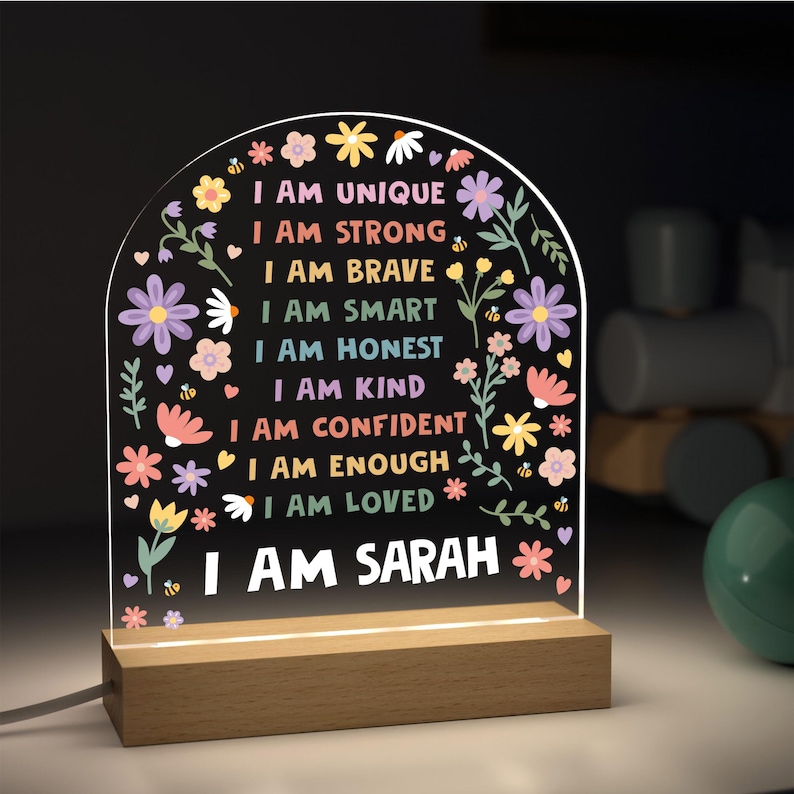May include: A decorative LED night light with a wooden base. The clear acrylic panel features colorful floral designs and the text "I AM UNIQUE, I AM STRONG, I AM BRAVE, I AM SMART, I AM HONEST, I AM KIND, I AM CONFIDENT, I AM ENOUGH, I AM LOVED, I AM SARAH."