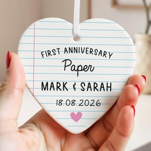 Puede incluir: Adorno de cer&aacute;mica en forma de coraz&oacute;n con dise&ntilde;o de papel rayado. El texto dice "FIRST ANNIVERSARY Paper MARK & SARAH 18.08.2026" con un coraz&oacute;n rosa. Se adjunta una cinta blanca para colgar.