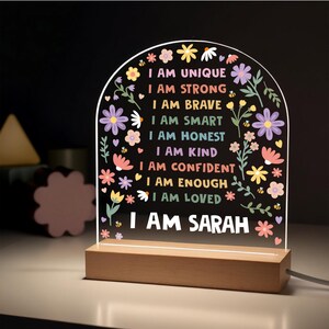 May include: A decorative LED night light with a wooden base. The clear acrylic panel features the text "I AM UNIQUE, I AM STRONG, I AM BRAVE, I AM SMART, I AM HONEST, I AM KIND, I AM CONFIDENT, I AM ENOUGH, I AM LOVED, I AM SARAH" surrounded by colorful floral designs.