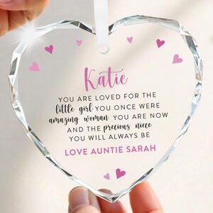 Puede incluir: Un adorno de cristal transparente en forma de coraz&oacute;n con una cinta blanca. El adorno tiene corazones rosas y el nombre "Katie" en escritura rosa. El texto dice: "YOU ARE LOVED FOR THE little girl...LOVE AUNTIE SARAH."