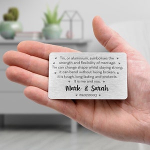 May include: A rectangular, silver-colored metal card held in a hand. The card has engraved text about marriage, including the names "Mark & Sarah" and the date "29.07.2003". The card's message is surrounded by small heart symbols.