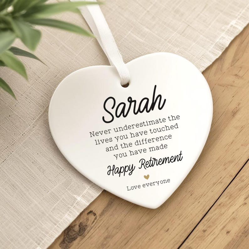 Op de afbeelding: Witte hartvormige keramische plaque met de naam "Sarah" in zwart schrift. De plaque bevat de tekst: "Onderschat nooit de levens die je hebt geraakt en het verschil dat je hebt gemaakt. Fijne pensioen. Love everyone."