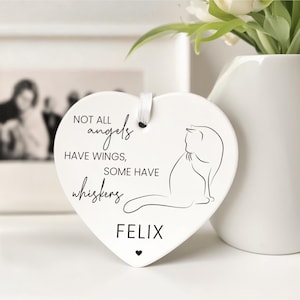 May include: A white heart-shaped ceramic ornament featuring a black cat outline and the text "NOT ALL angels HAVE WINGS, SOME HAVE whiskers FELIX". A small black heart is at the bottom. A white ribbon is attached for hanging.