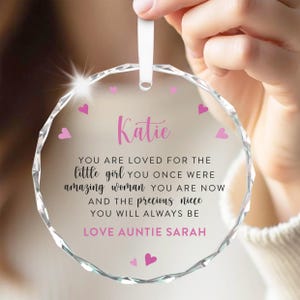 Puede incluir: Un adorno de cristal transparente y facetado con una cinta blanca. El adorno tiene corazones rosas y el nombre "Katie" en escritura rosa. El texto dice "YOU ARE LOVED FOR THE little girl YOU ONCE WERE...LOVE AUNTIE SARAH."