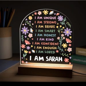May include: A decorative LED night light with a wooden base. The acrylic panel features colorful floral designs and the phrases "I AM UNIQUE" to "I AM LOVED", and "I AM SARAH" in white text. The light is on.