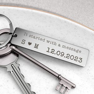 May include: Silver rectangular keychain with the engraved text "It started with a message..." and the date 12.09.2023. The keychain also features the initials "S &hearts; M". Two keys are visible next to the keychain.