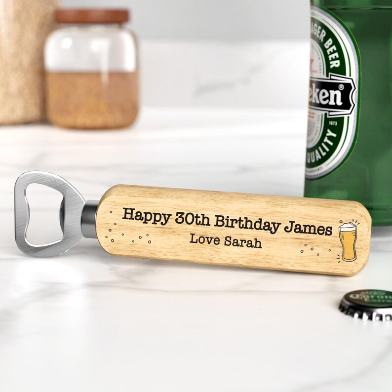 Puede incluir: Un abridor de botellas de madera con un abridor de metal plateado. El mango est&aacute; grabado con "Happy 30th Birthday James Love Sarah" y un gr&aacute;fico de vaso de cerveza. Una botella de cerveza verde est&aacute; en el fondo.