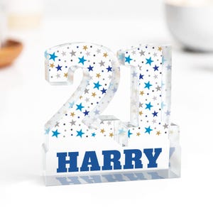 Puede incluir: Un n&uacute;mero 21 de acr&iacute;lico transparente con un patr&oacute;n de estrellas azules, doradas y plateadas. La base muestra el nombre "HARRY" en letras azules. Un adorno decorativo.