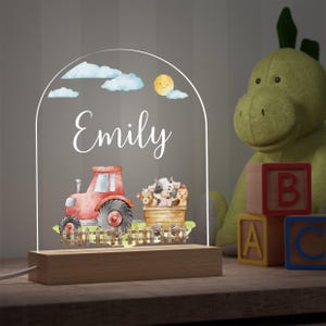 May include: A personalised night light with the name "Emily" in white script. The design features a red tractor, a wooden bucket with farm animals, and a yellow sun with clouds. The light sits on a wooden base.