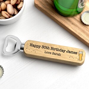 Puede incluir: Un abridor de botellas de madera con cabezal de acero inoxidable. El abridor est&aacute; grabado con "Happy 30th Birthday James Love Sarah" y un icono de vaso de cerveza. El abridor est&aacute; sobre una superficie blanca.