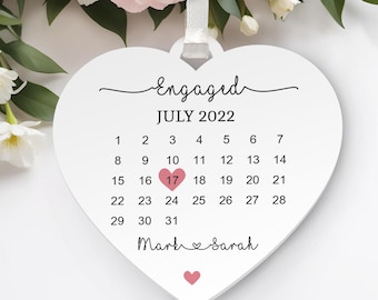 以下が含まれることがあります： 「Engaged」の文字と2022年7月のカレンダーが描かれた、白いハート型の婚約オーナメント。17日はピンクのハートで強調されています。「Mark & Sarah」という名前がカレンダーの下に書かれており、ピンクのハートが付いています。