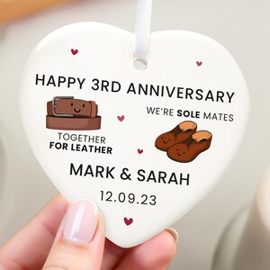 May include: A heart-shaped ceramic ornament with a white ribbon. It features the text "HAPPY 3RD ANNIVERSARY" and illustrations of a belt and shoes. The text "WE'RE SOLE MATES" and "TOGETHER FOR LEATHER" are also included, along with a date.