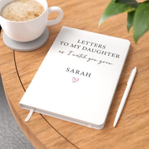 May include: A white hardback journal featuring the black printed text "LETTERS TO MY DAUGHTER as I watch you grow" and the name "SARAH" above a small pink heart icon. The journal is placed beside a white pen and a white mug containing coffee.