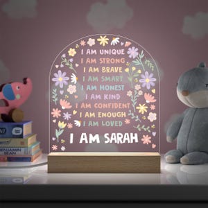 May include: A decorative night light with a wooden base and a clear acrylic arch. The arch features floral designs and the text "I AM UNIQUE, I AM STRONG, I AM BRAVE, I AM SMART, I AM HONEST, I AM KIND, I AM CONFIDENT, I AM ENOUGH, I AM LOVED, I AM SARAH."