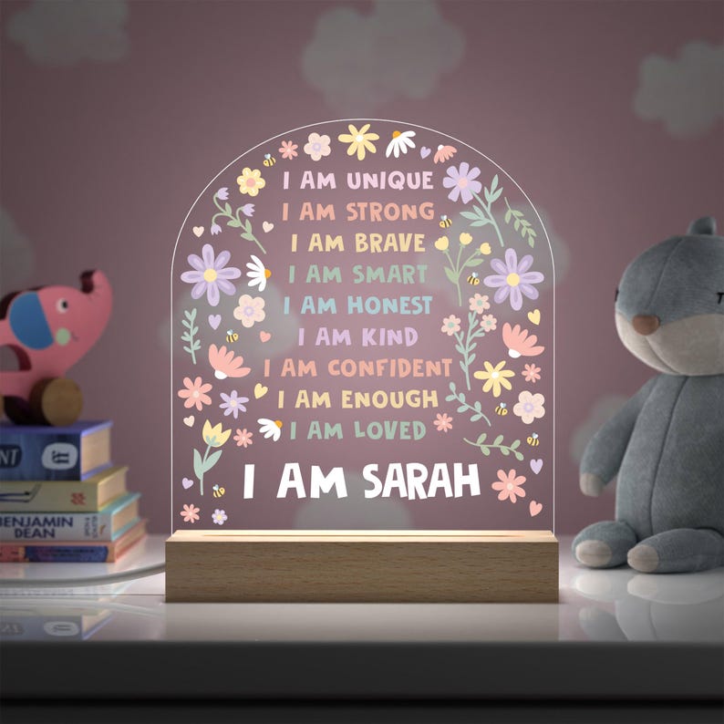 May include: A decorative night light with a wooden base and a clear acrylic arch. The arch features floral designs and the text "I AM UNIQUE, I AM STRONG, I AM BRAVE, I AM SMART, I AM HONEST, I AM KIND, I AM CONFIDENT, I AM ENOUGH, I AM LOVED, I AM SARAH."